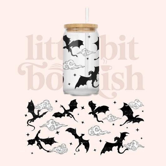 A clear glass can-style tumbler with a bamboo lid and white straw, printed with black flying dragons and white clouds on a pale pink background. A matching sheet of the same dragons-and-cloud designs appears below, and a faint 'little bit bookish' watermark is visible across the image.
