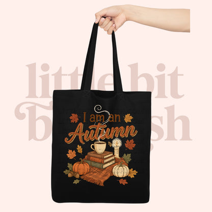 A black tote bag with a hand holding the top handle, featuring a fall-themed graphic that reads 'I am an Autumn' with pumpkins, leaves, books, and a mug. The background is a pale peach-pink with a faint watermark.