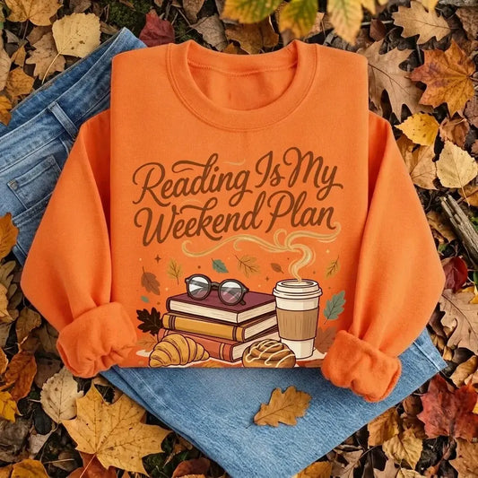 An orange crewneck sweatshirt with a cozy autumn-themed graphic reading 'Reading Is My Weekend Plan' in brown script. The graphic features books, reading glasses, a coffee cup, and pastries.