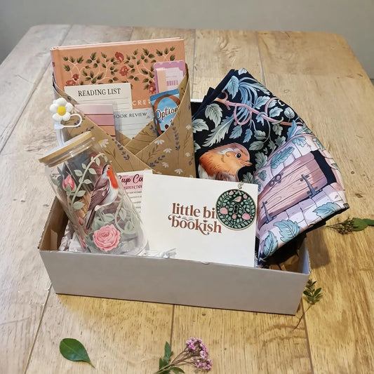 A curated bookish gift box set on a light wood tabletop, containing a floral-patterned reading list card, a small jar with a floral label, a white card reading 'little bit bookish,' folded patterned fabric, and other coordinated stationery or gift items.
