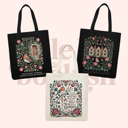 Three decorative canvas tote bags shown on a pale pink background: two black bags with illustrated garden and house designs, and one cream bag with a floral and bird circular design and text reading 'The Secret Garden' and 'Misselthwaite Manor.'.