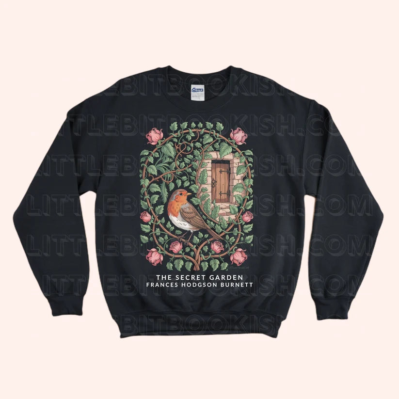 A black crewneck sweatshirt featuring a detailed graphic of a robin bird in a secret garden with pink flowers and a wooden door, with the text 'THE SECRET GARDEN FRANCES HODGSON BURNETT' printed below. The sweatshirt is displayed flat against a light beige background with a faint watermark pattern.