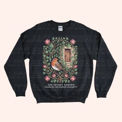 A black crewneck sweatshirt featuring a detailed graphic of a robin bird in a secret garden with pink flowers and a wooden door, with the text 'THE SECRET GARDEN FRANCES HODGSON BURNETT' printed below. The sweatshirt is displayed flat against a light beige background with a faint watermark pattern.