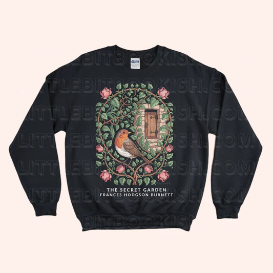 A black crewneck sweatshirt featuring a detailed graphic of a robin bird in a secret garden with pink flowers and a wooden door, with the text 'THE SECRET GARDEN FRANCES HODGSON BURNETT' printed below. The sweatshirt is displayed flat against a light beige background with a faint watermark pattern.